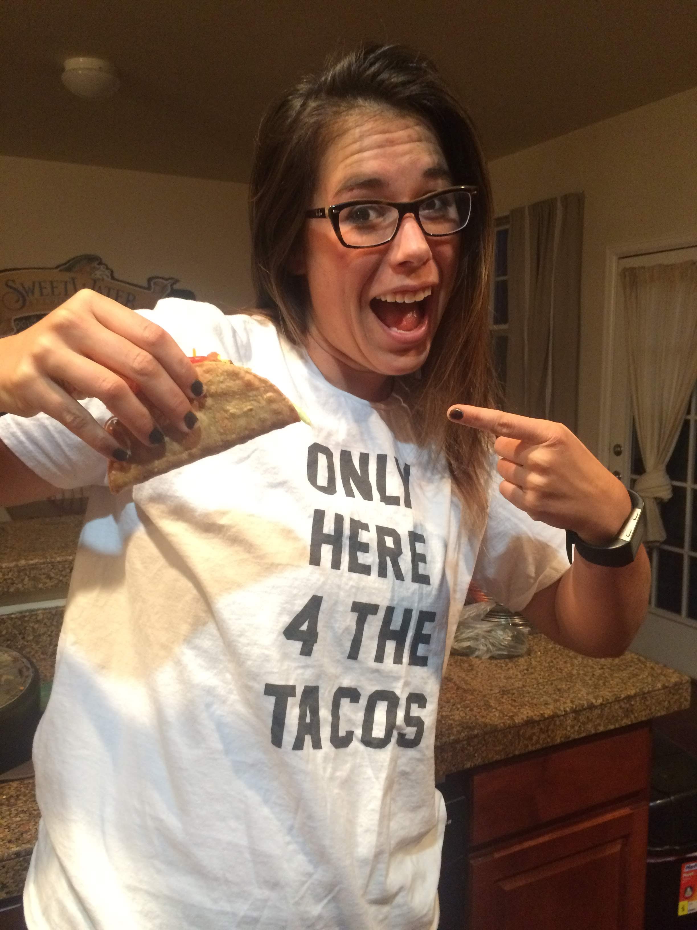 A woman wearing a white t-shirt with the text 'ONLY HERE 4 THE TACOS' is smiling and holding a taco, pointing at it.